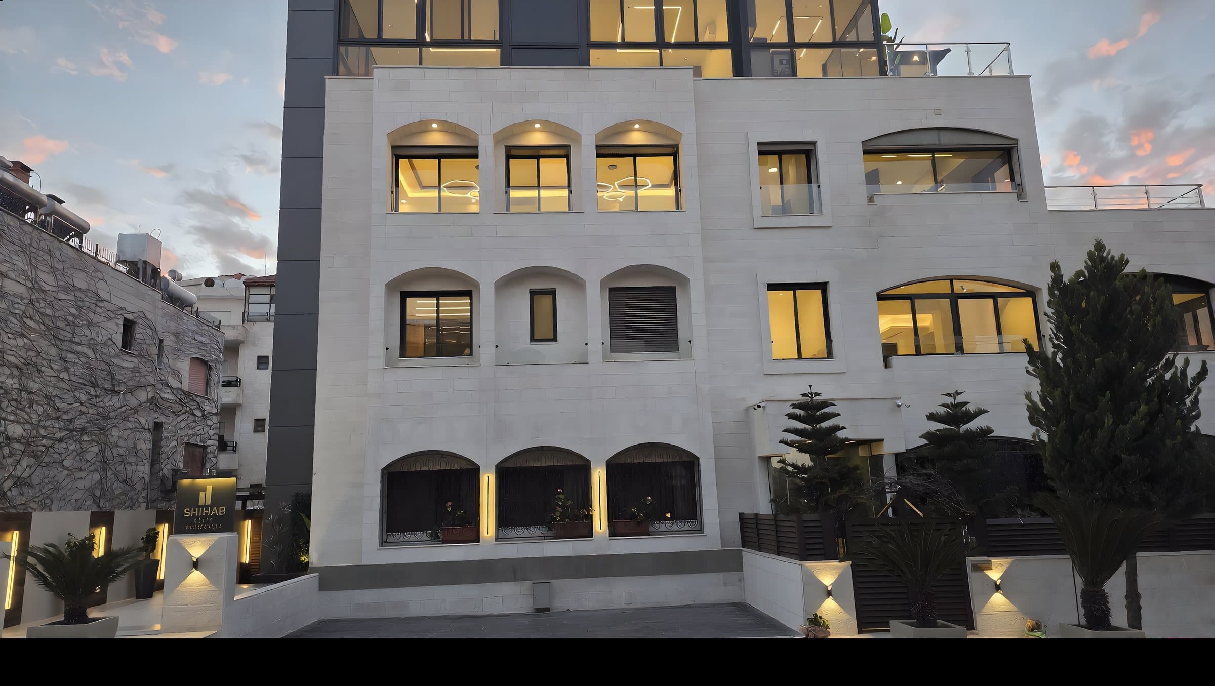 SHIHAB Elite Residences stone facade at dusk, warmly lit windows set back from the street with mature planting