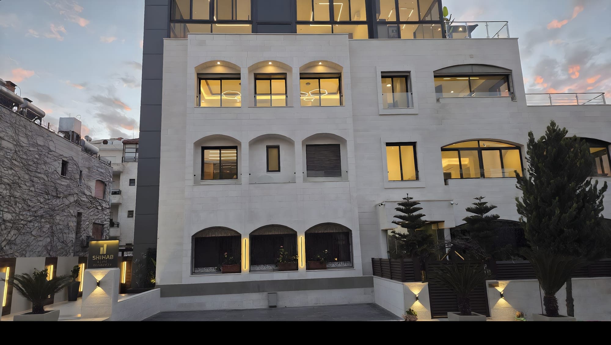 SHIHAB Elite Residences stone facade at dusk, warmly lit windows set back from the street with mature planting